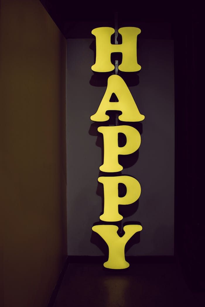 about-img Illuminated yellow 'HAPPY' sign displayed vertically in a dimly lit hallway setting, evoking a playful mood.